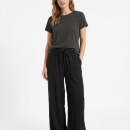 Femeie purtând un tricou gri și pantaloni negri, stând în fața unui fundal alb.